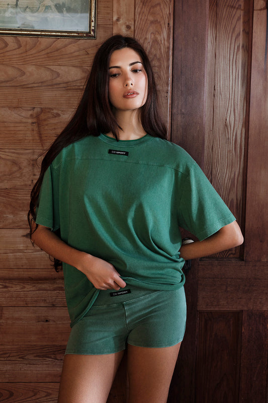 Woman wearing a green t-shirt and shorts set against a wooden wall. |M1