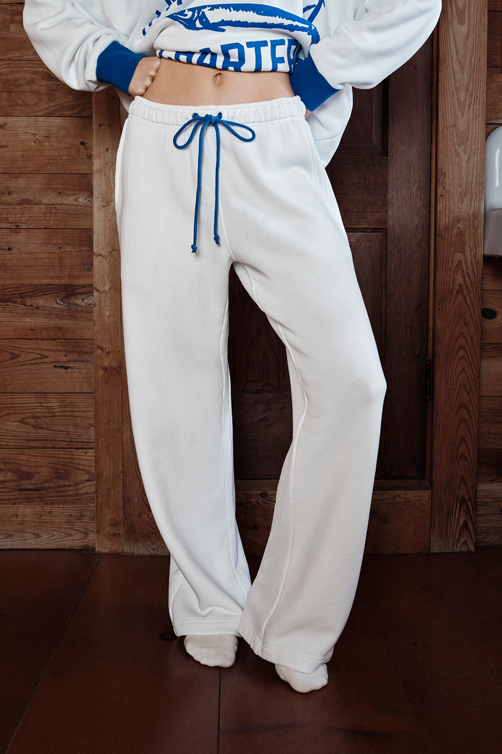 Person wearing a white sweatshirt with blue accents and white sweatpants against a wooden background |M1
